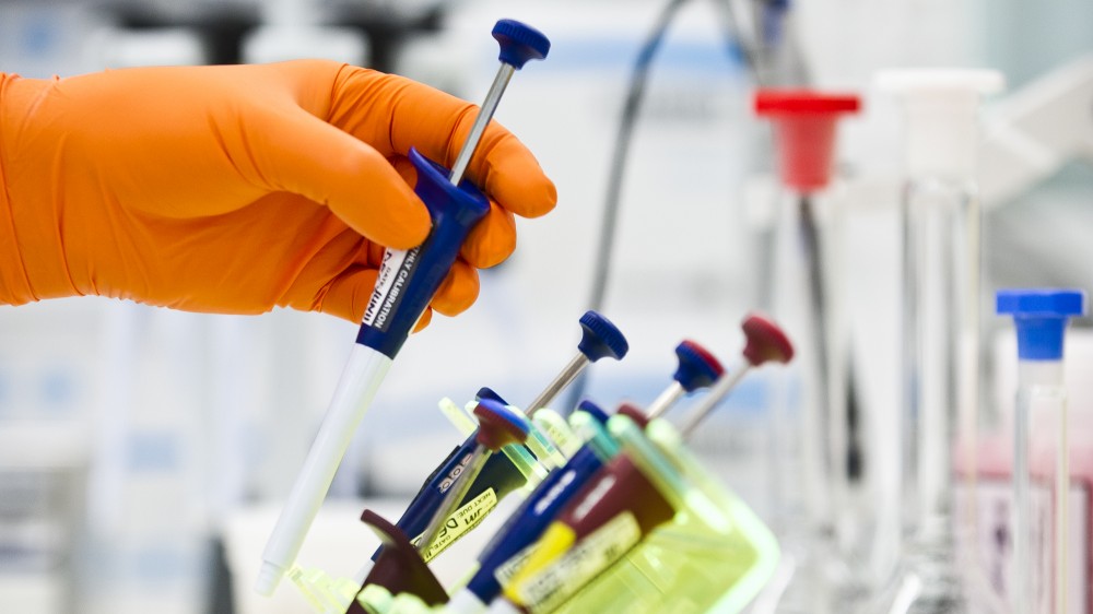 An orange gloved hand holds a pipette above a row of other pipettes on a lab bench.