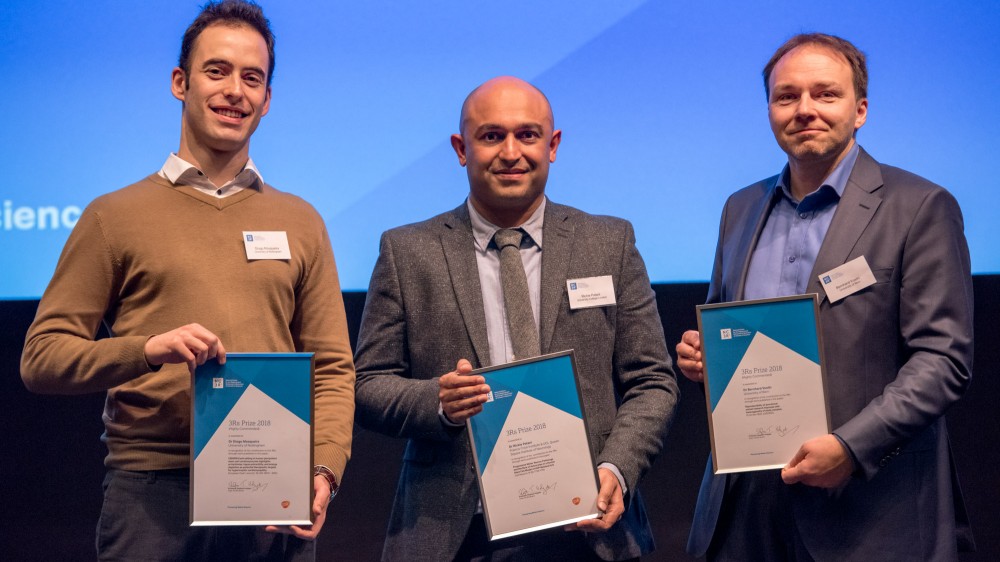 The three winners of the 2018 3Rs prize. Left to right: Dr Diogo Mosqueria, highly commended. Dr Rickie Patani, winner. Dr Bernhard Voelkl. All three men are stood on stage in front of a presentation screen holding their prize certificates in front of them.