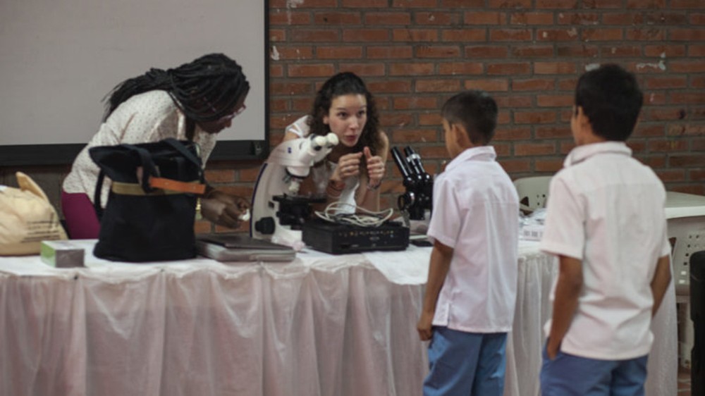 Dr María Duque-Correa in conversation with two young students at a table with scientific equipment