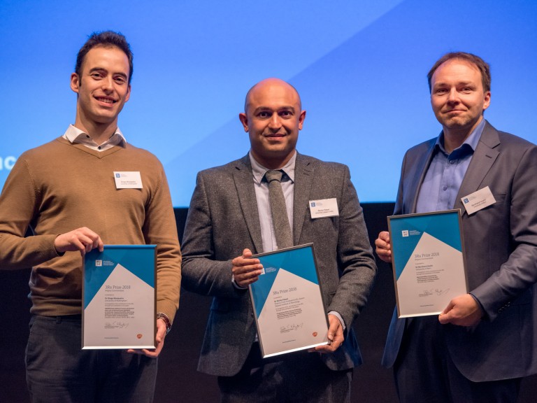 The three winners of the 2018 3Rs prize. Left to right: Dr Diogo Mosqueria, highly commended. Dr Rickie Patani, winner. Dr Bernhard Voelkl. All three men are stood on stage in front of a presentation screen holding their prize certificates in front of them.