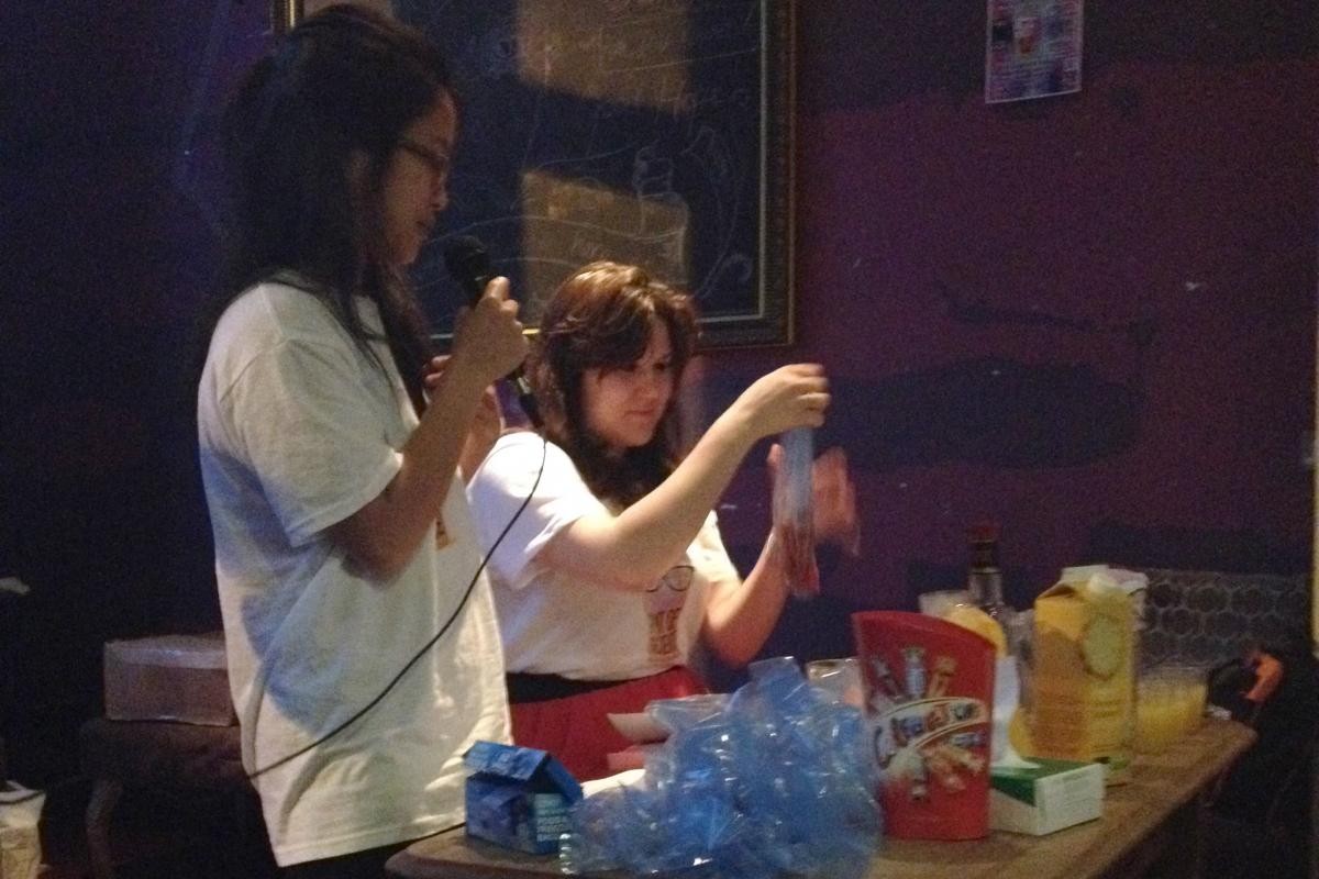 Two ladies, one sitting down and one standing with a microphone behind a table with various food items on it for 3Rs-related educational fun