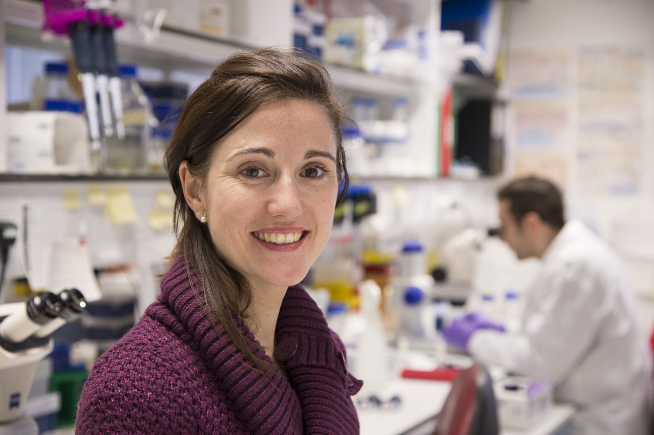 Dr Dr Meritxell Huch, winner of the 2013 3Rs prize. She is in a laboratory setting and you can see a researcher in a white coat and purple gloves looking down a microscope in the background.